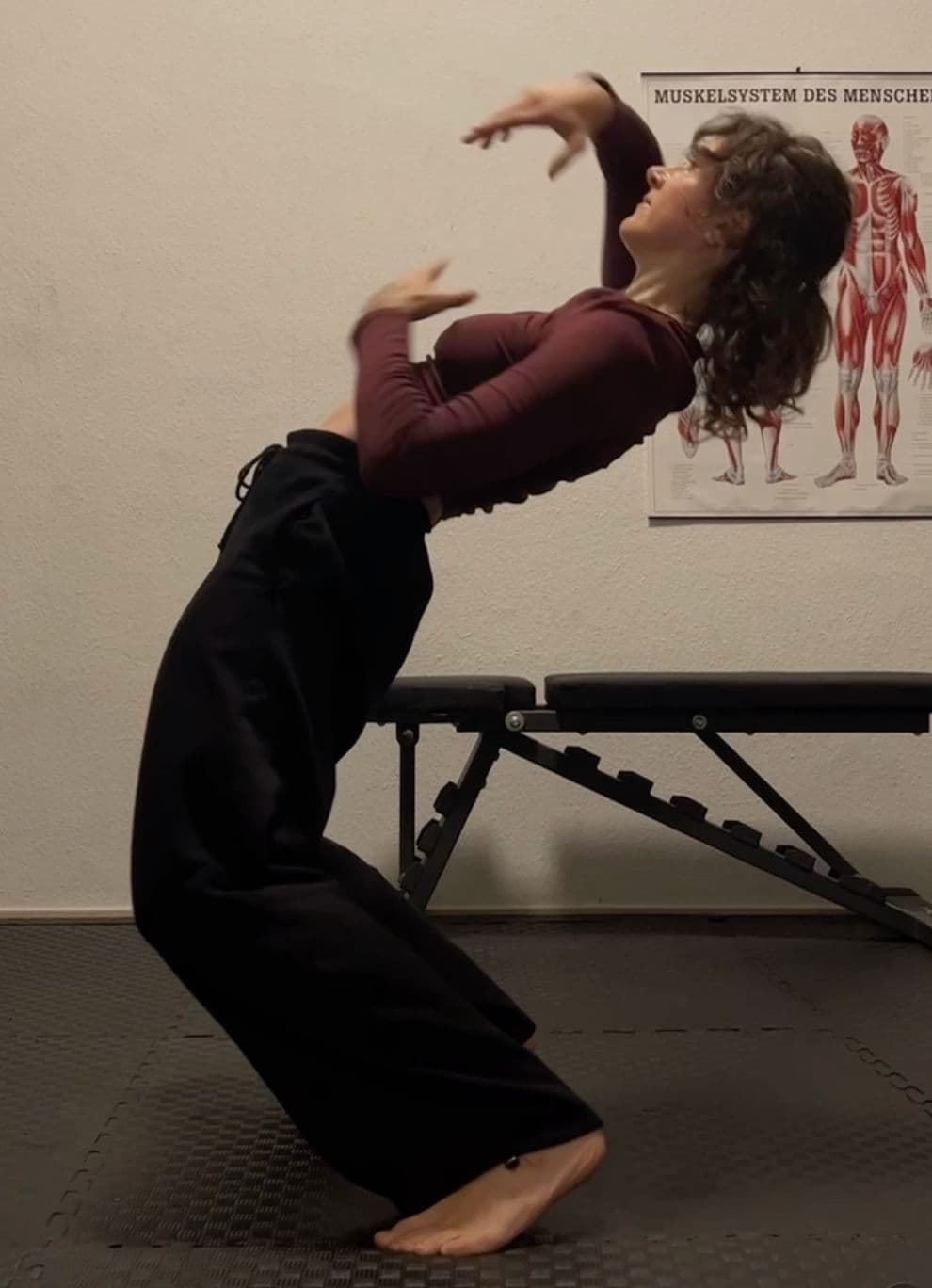 Clara in a deep backbend squat in a dimly lit studio, arms stretched overhead with a human anatomy chart on the wall behind her.