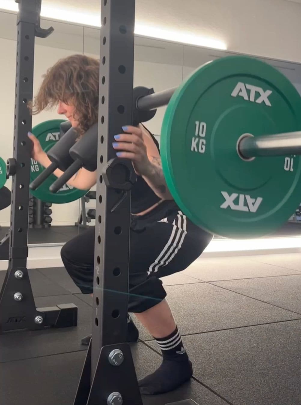 Back view of Clara performing a barbell back squat with green weight plates in a gym.