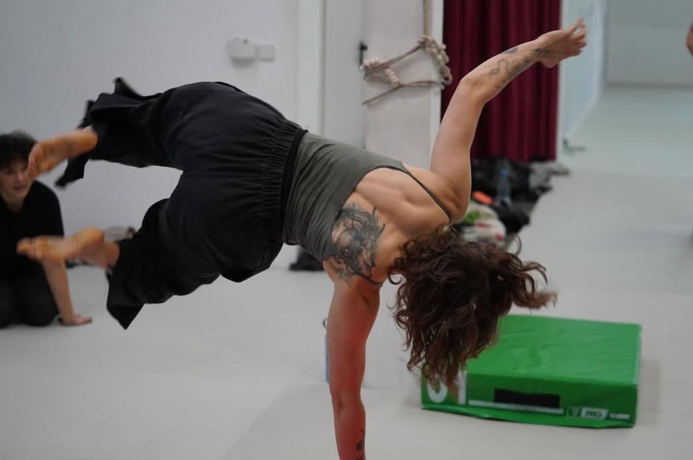 Clara mid-handstand in a movement studio, one leg extended overhead and loose clothing flowing, hands pressed into a green block.