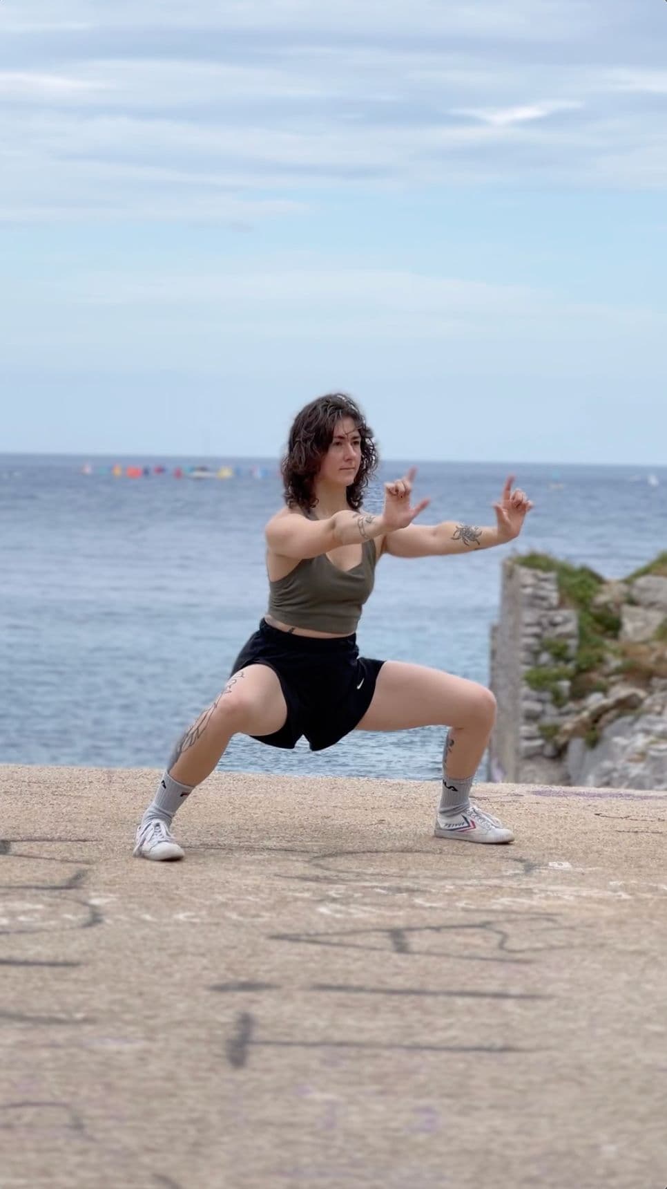 Clara in a low horse stance on a concrete platform by the sea, arms extended, grey ocean and overcast sky behind her.
