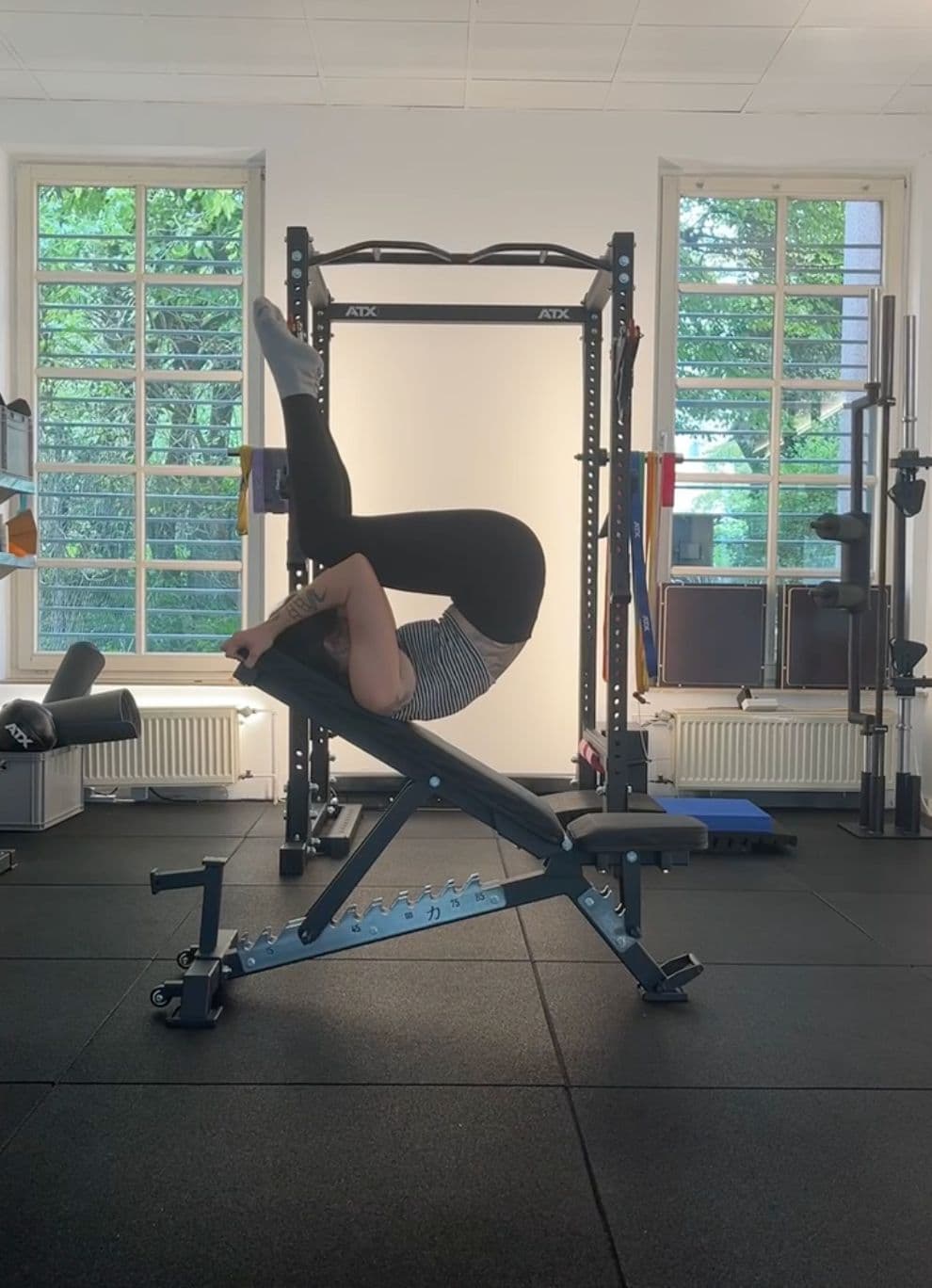 Clara upside-down in a controlled inversion over a bench in a light-filled studio, legs raised and curved above her.