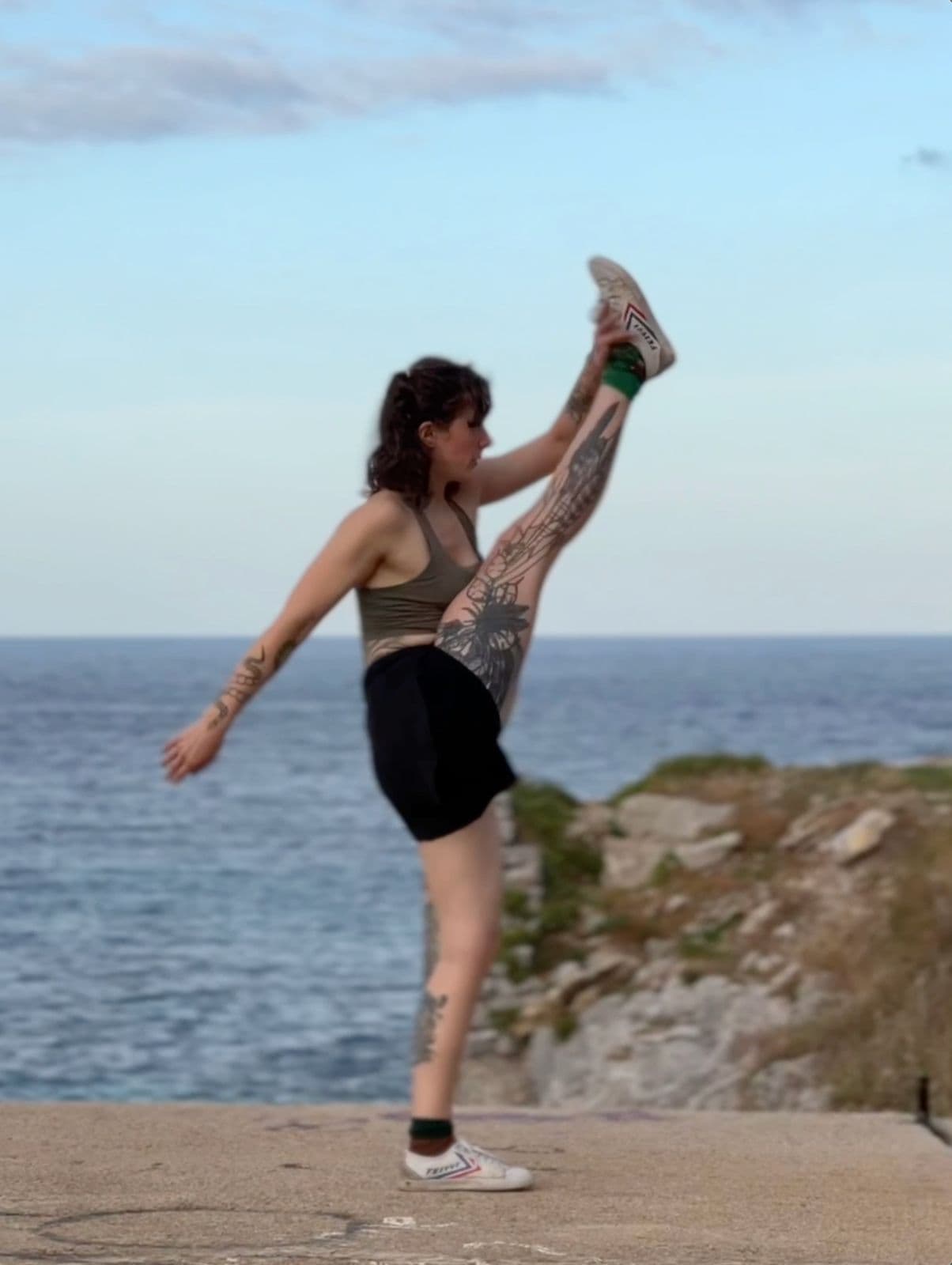Clara standing with one leg raised high and hand clasping her foot, ocean behind her and soft evening sky.