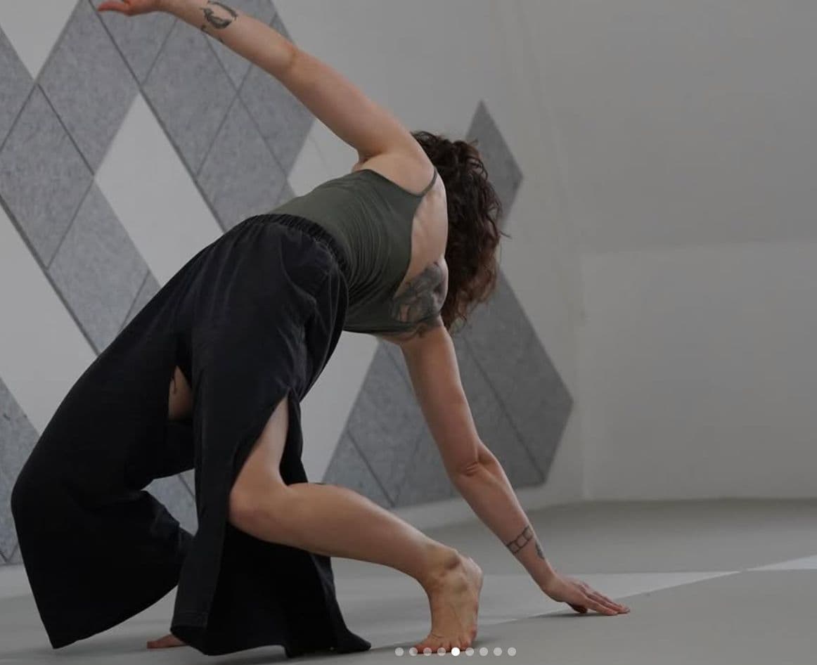 Clara in a deep low lunge reaching for the floor, photographed against a wall of diamond-patterned tiles in warm directional light.