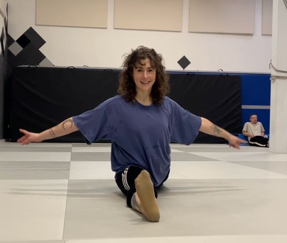 Clara seated cross-legged on a mat in a martial arts studio, arms spread wide, smiling warmly at the camera.