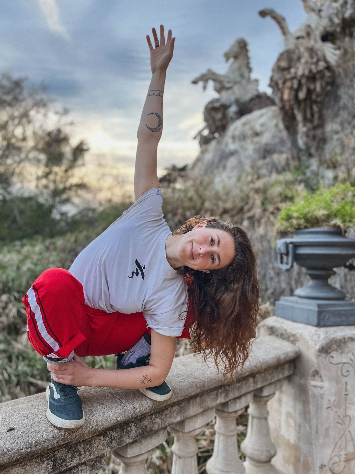 Clara balancing in a deep side bend on a stone balustrade in a park at dusk, one arm reaching toward the sky.